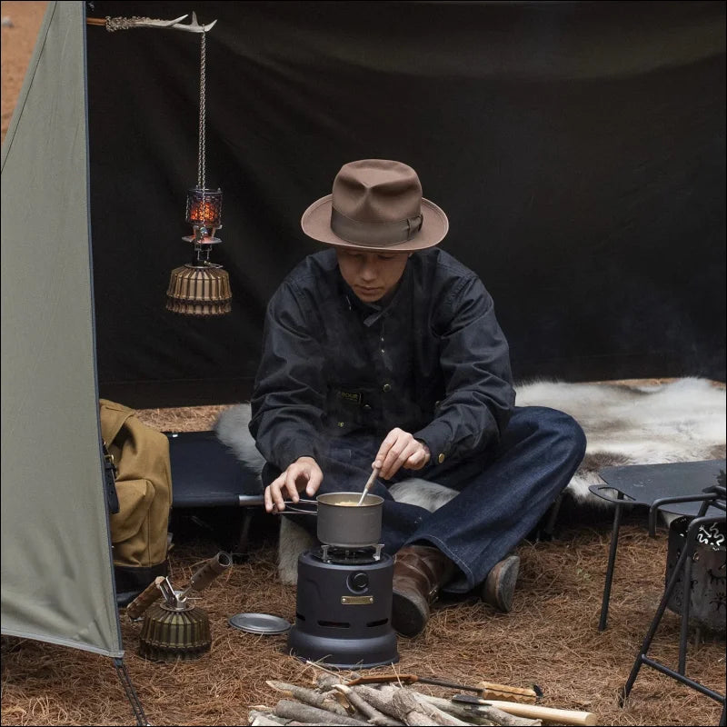 Mit hut und jacke bekleidete person, die im schneidersitz auf einem tragbaren kocher in einer campingumgebung kocht.