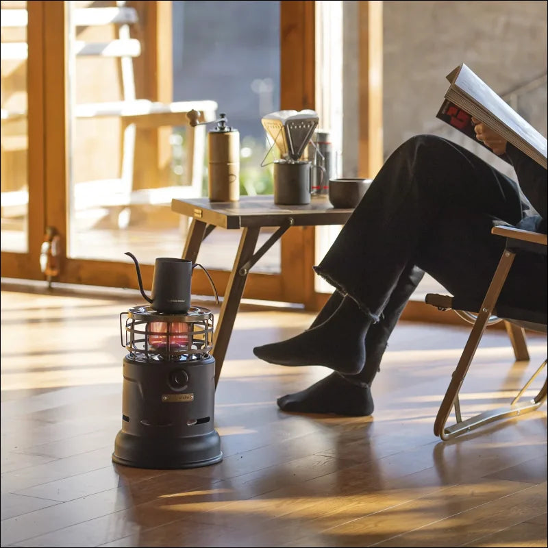 Auf einem stuhl sitzende person, die liest, mit einem tragbaren heizgerät zu ihren füßen und kaffeezubehör auf einem tisch in der nähe.